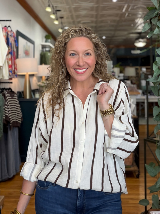 A smiling woman with curly blonde hair stands in a boutique wearing a cream-colored button-down shirt with dark brown vertical stripes and blue jeans.