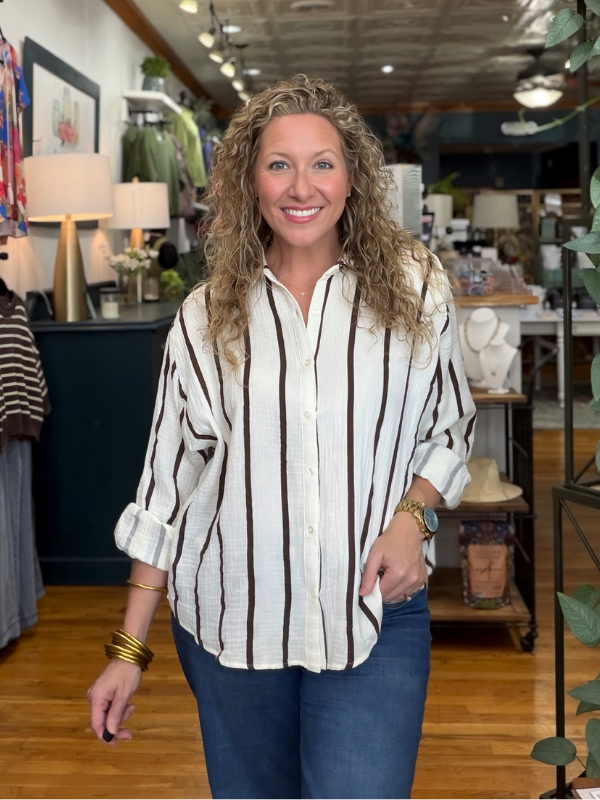 A smiling woman with curly blonde hair stands in a boutique wearing a white button-up shirt with vertical brown stripes and blue jeans.