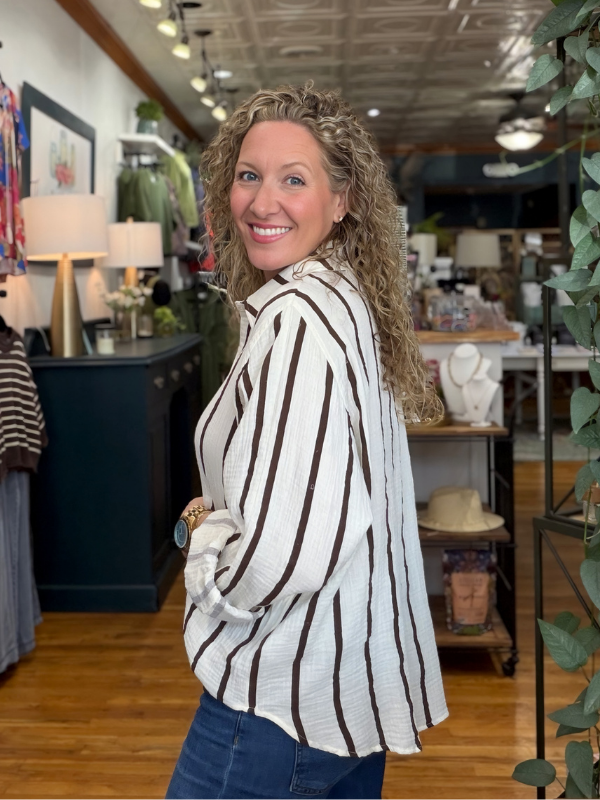 A smiling woman with curly hair wearing a white button-up shirt with dark brown vertical stripes and blue jeans, standing inside a retail boutique.