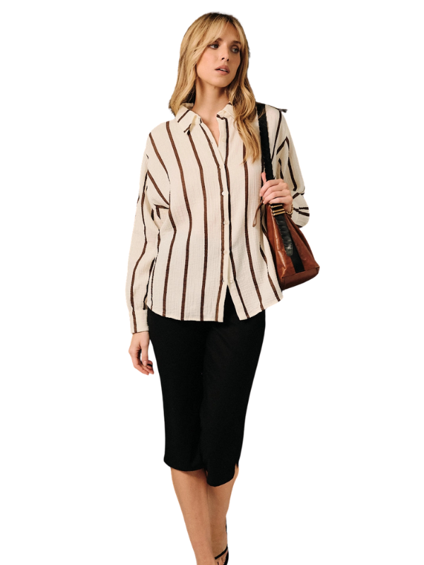 A woman modeling a cream-colored long-sleeve button-up shirt with vertical brown stripes, paired with black capri pants and a brown shoulder bag, set against a plain white background.