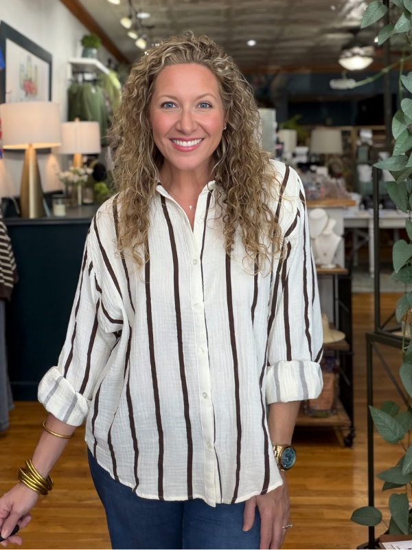 A smiling woman with curly blonde hair wearing a cream-colored linen button-up shirt with dark brown vertical stripes, paired with dark blue jeans, standing inside a furniture store.