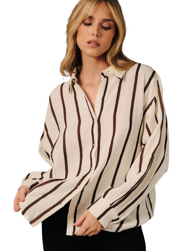 A woman wearing a cream-colored long-sleeve button-up shirt with vertical dark brown stripes, paired with black pants, against a plain white background.
