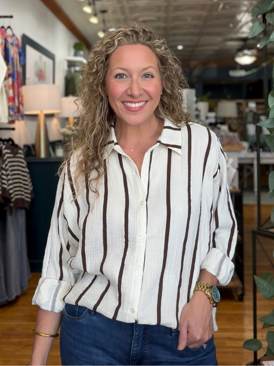 A smiling woman with curly blonde hair stands in a boutique wearing a cream-colored button-down shirt with dark brown vertical stripes and blue jeans.