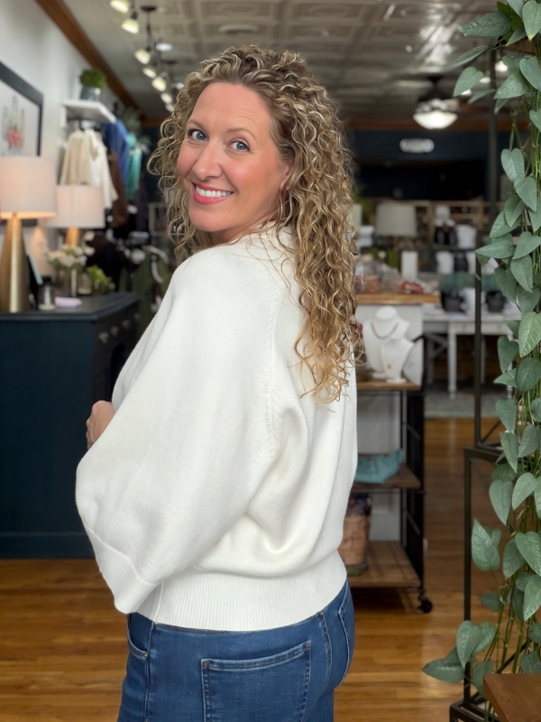 A woman with curly blonde hair wearing a cream-colored knit sweater and blue jeans, posing inside a retail store.