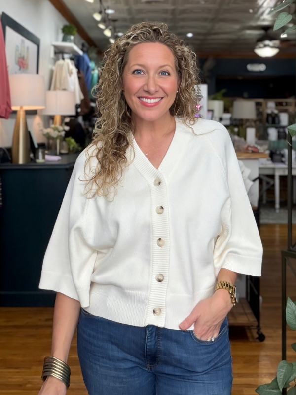 A smiling woman with curly blonde hair stands in a boutique wearing a cream-colored knit cardigan with three-quarter sleeves and front buttons, paired with blue jeans.