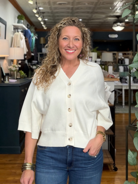 A smiling woman with curly blonde hair stands in a boutique wearing a cream-colored knit cardigan with button-front detailing and blue jeans.
