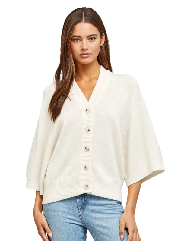 A woman modeling a cream-colored knit poncho cardigan with wide three-quarter sleeves and a button-down front, paired with light blue jeans against a plain white background.