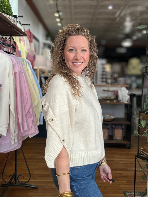 Woman in a white sweater standing in a store with clothing racks and products in the background