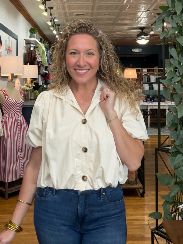 A smiling woman with curly blonde hair wearing a cream puff-sleeve button-up shirt and dark blue jeans, standing inside a boutique store.