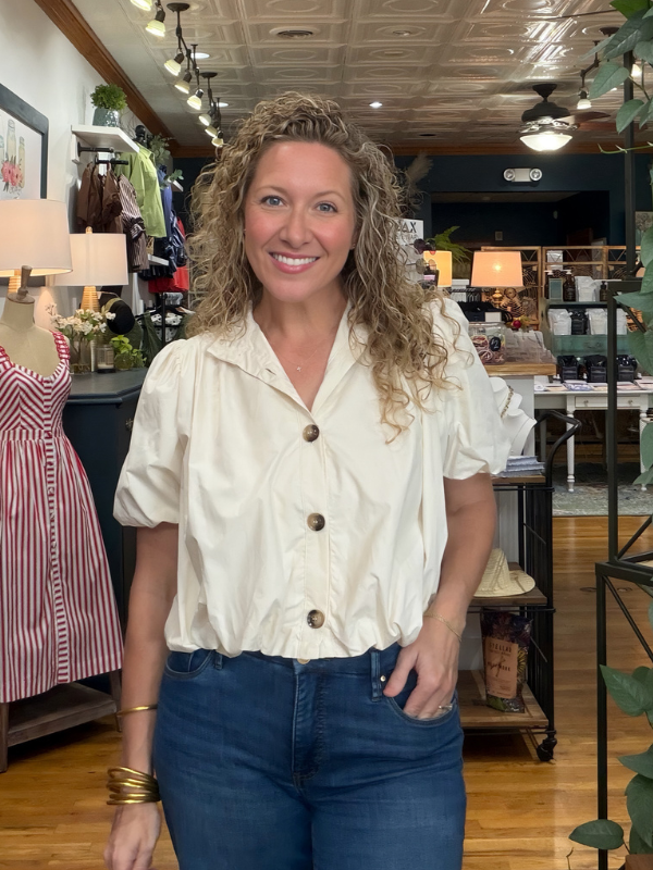 A smiling woman with curly blonde hair stands inside a boutique shop wearing a cream puff-sleeve button-up blouse and blue jeans.
