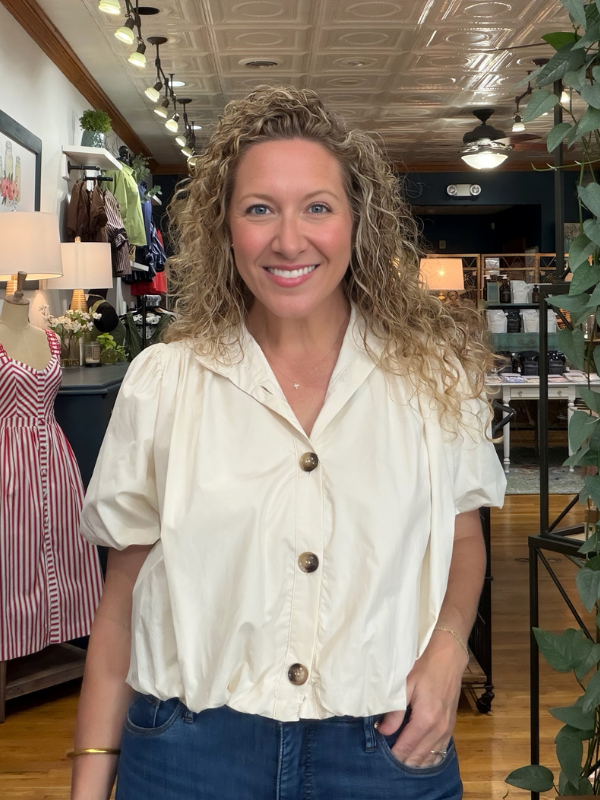A smiling woman with curly blonde hair wearing a cream-colored short-sleeve button-up blouse and blue jeans, standing inside a retail store.