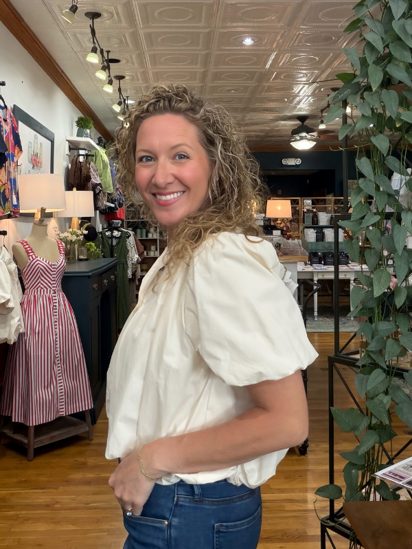 A smiling woman with curly hair stands inside a boutique shop, wearing a cream puff-sleeve blouse and blue jeans.