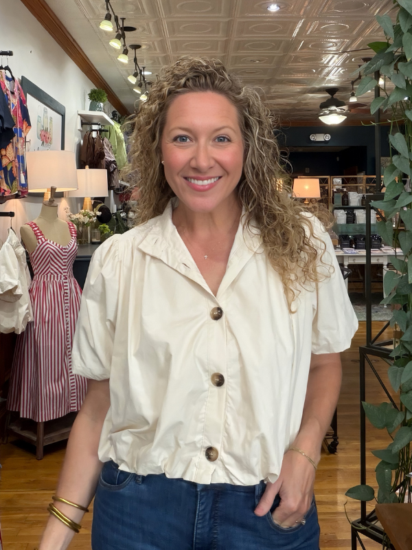 A smiling woman with curly blonde hair wearing a cream-colored short-sleeve button-up blouse and blue jeans, standing inside a retail store.