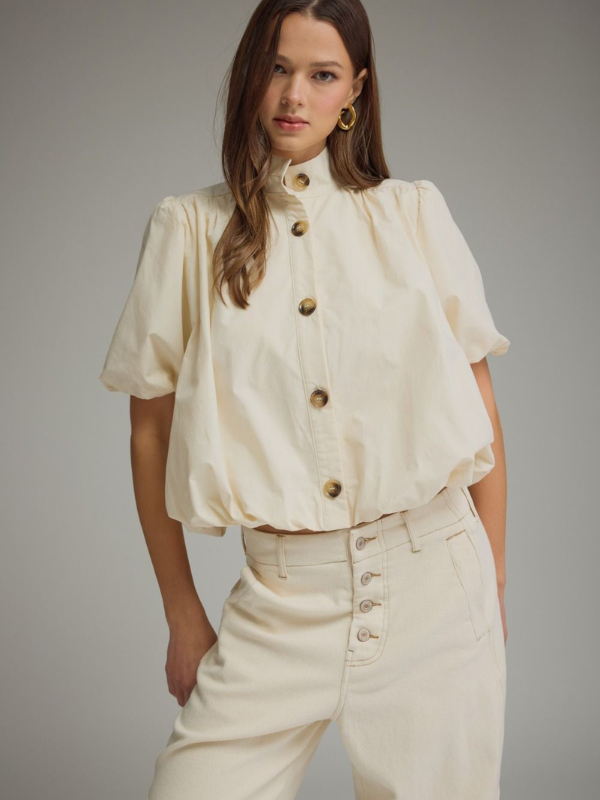 Woman wearing a cream-colored blouse with puff sleeves and high-waisted pants against a gray background