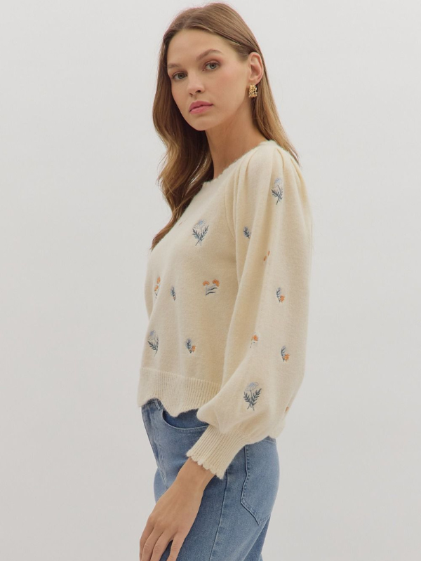 Woman wearing a cream sweater with small embroidered designs on a plain background