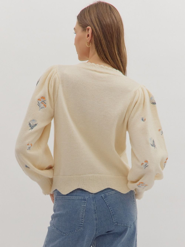 Person wearing a beige sweater with small embroidered designs on a plain background