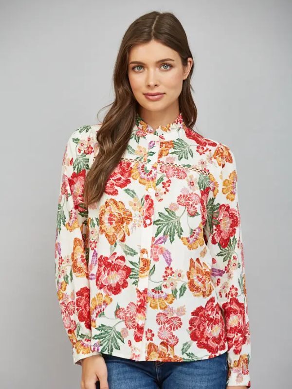 A woman modeling a long-sleeve high-neck blouse with a vibrant multicolor floral print on a cream background, paired with blue jeans, against a solid gray studio background.