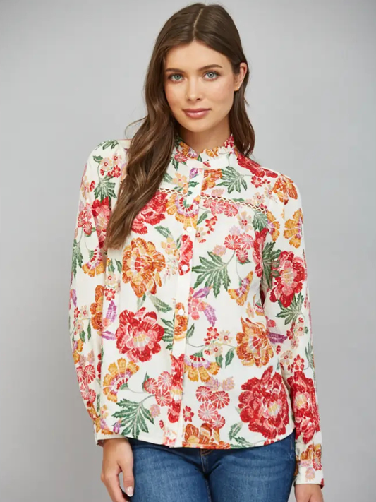 A woman modeling a long-sleeve high-neck blouse with a vibrant multicolor floral print on a cream background, paired with blue jeans, against a solid gray studio background.