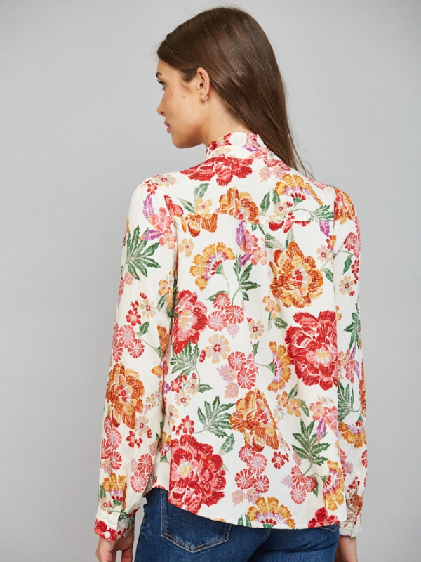 A woman shown from the back wearing a long-sleeve floral blouse with a white base and red, orange, pink, and green floral print, paired with blue jeans against a light gray studio background.