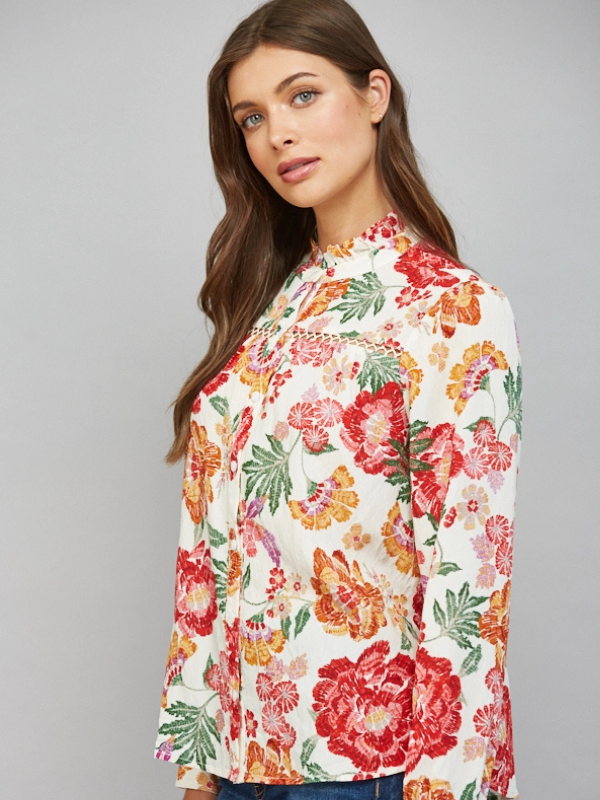 A woman wearing a long-sleeve floral blouse with a high mock neck, featuring a vibrant red, orange, and green floral pattern on an off-white background, photographed against a solid gray studio backdrop.