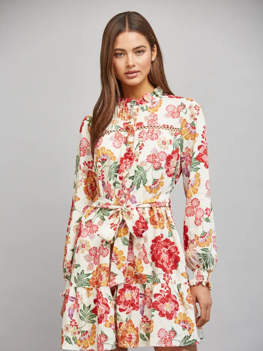 A woman modeling a long-sleeve floral dress with a tie waist, featuring a cream base with red, pink, and green flowers, against a solid gray studio background.