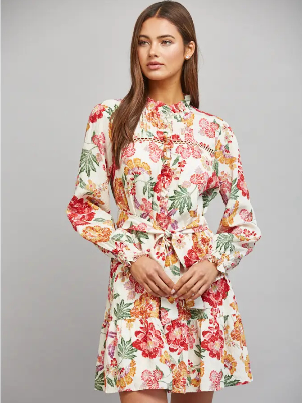 A woman modeling a long-sleeve floral dress with a tie waist, photographed against a plain light gray studio background.