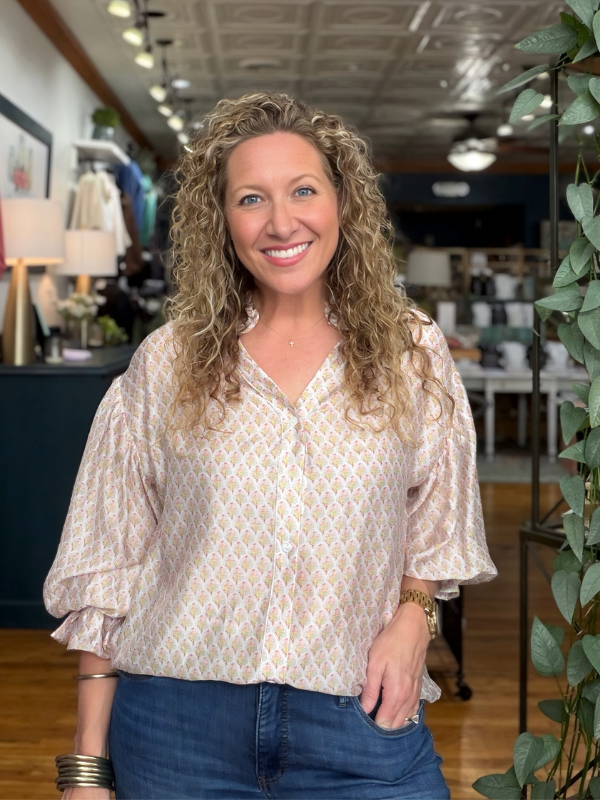 A smiling woman with curly blonde hair wearing a light beige and white patterned blouse with blue jeans, standing inside a boutique store.