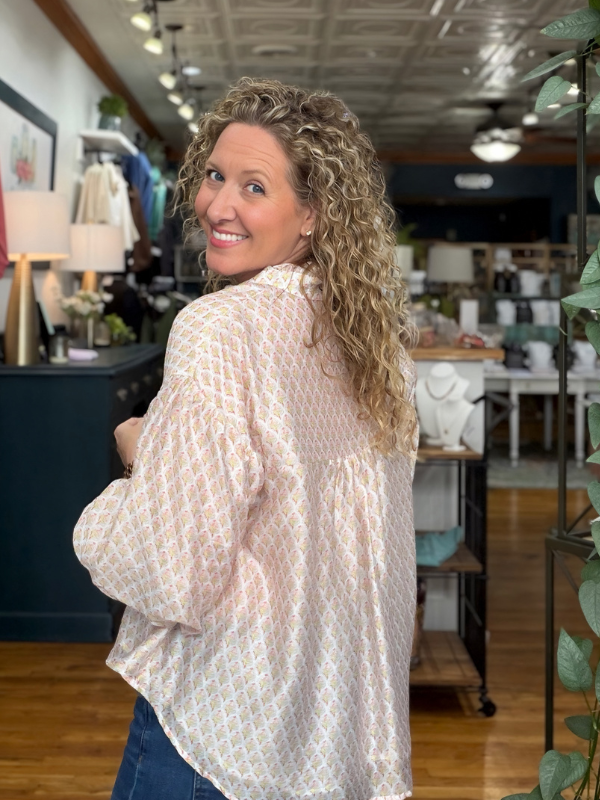 A woman with curly blonde hair wearing a light pink and white patterned long-sleeve blouse, photographed from behind inside a clothing boutique.