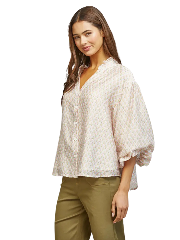 A woman modeling a loose, short-sleeved button-up blouse with a delicate geometric pattern in light pink and beige, paired with olive green pants, against a solid white background.