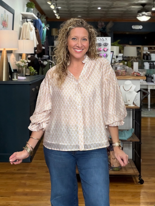 A smiling woman with curly blonde hair stands in a boutique shop wearing a light beige and white patterned button-down blouse with three-quarter ruffled sleeves, paired with blue jeans.
