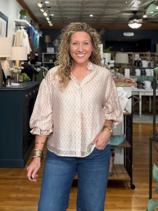 A smiling woman with curly blonde hair stands in a retail store wearing a light beige and white patterned blouse with ruffled sleeves and blue jeans.
