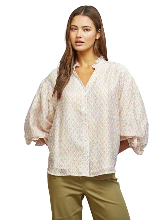A woman modeling a loose, short-sleeved button-up blouse with a delicate beige and white geometric pattern and voluminous ruffled sleeves, paired with olive green pants against a plain white background.