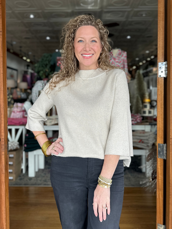 Woman standing in a store with a neutral expression, wearing a light gray sweater and dark jeans.