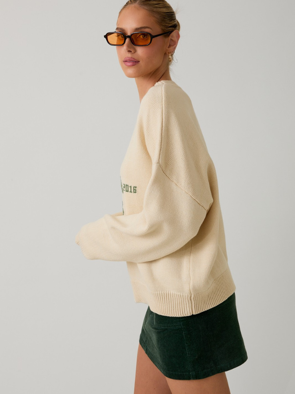 Woman wearing a beige sweater with text and a green skirt on a plain background