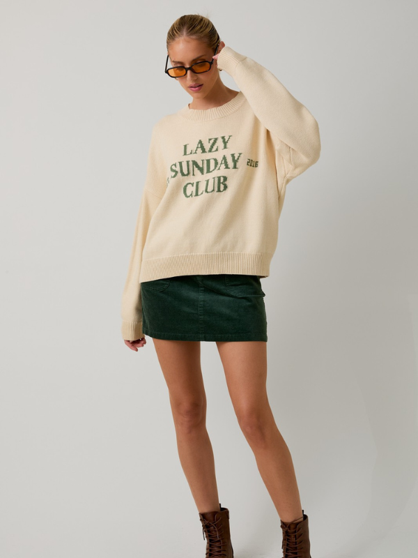 Woman wearing a beige sweater with 'Lazy Sunday Club' text, green skirt, and brown boots on a plain background
