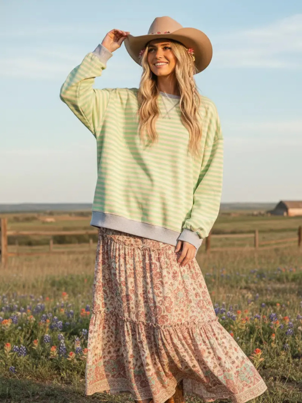 Woman wearing a light green striped sweater and patterned skirt standing in a field with a cowboy hat.