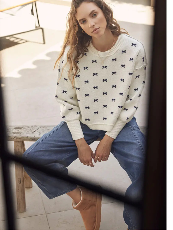 Woman wearing a white sweater with black bow patterns and blue jeans, sitting on a wooden bench.