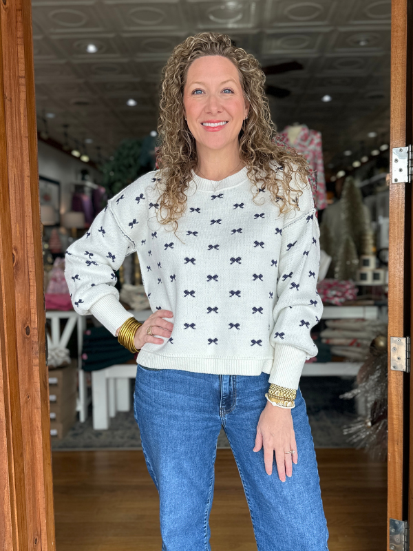 Woman wearing a white sweater with black bow pattern and blue jeans in a store setting.