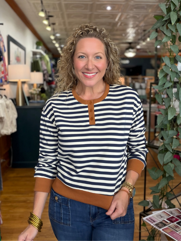 A smiling woman with curly blonde hair stands in a boutique wearing a navy and white horizontally striped long-sleeve henley top with brown trim, paired with blue jeans.