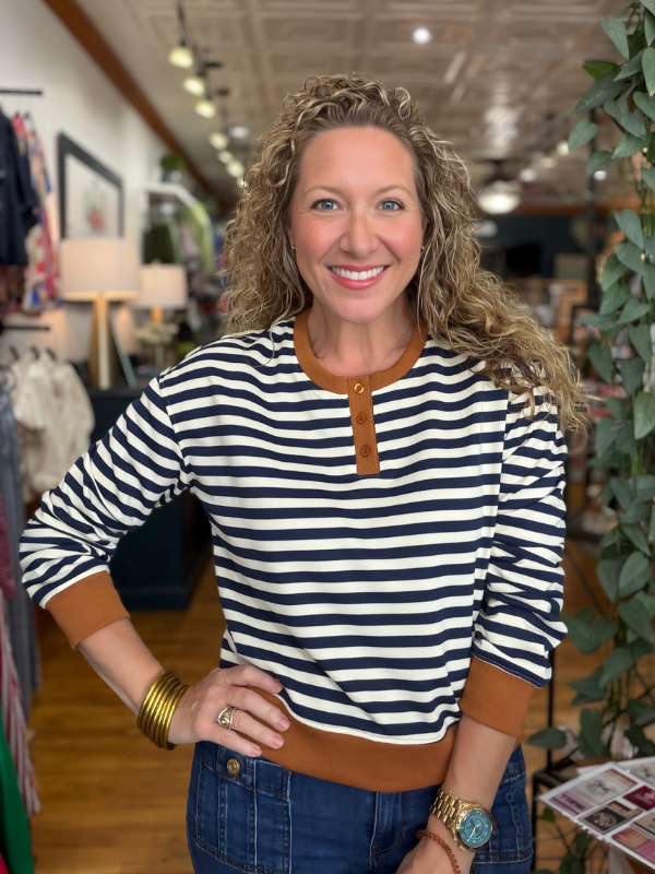 A smiling woman with curly blonde hair stands in a boutique wearing a navy and white horizontally striped henley top with brown trim, paired with blue jeans.