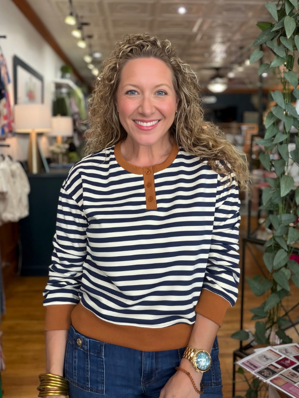 A smiling woman with curly blonde hair stands in a boutique wearing a navy and white horizontally striped henley top with brown trim, paired with blue jeans.