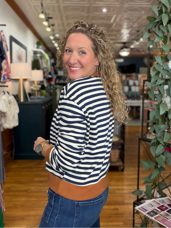 A woman with curly blonde hair wearing a navy and white striped long-sleeve top with a brown waistband and blue jeans, standing in a retail store.