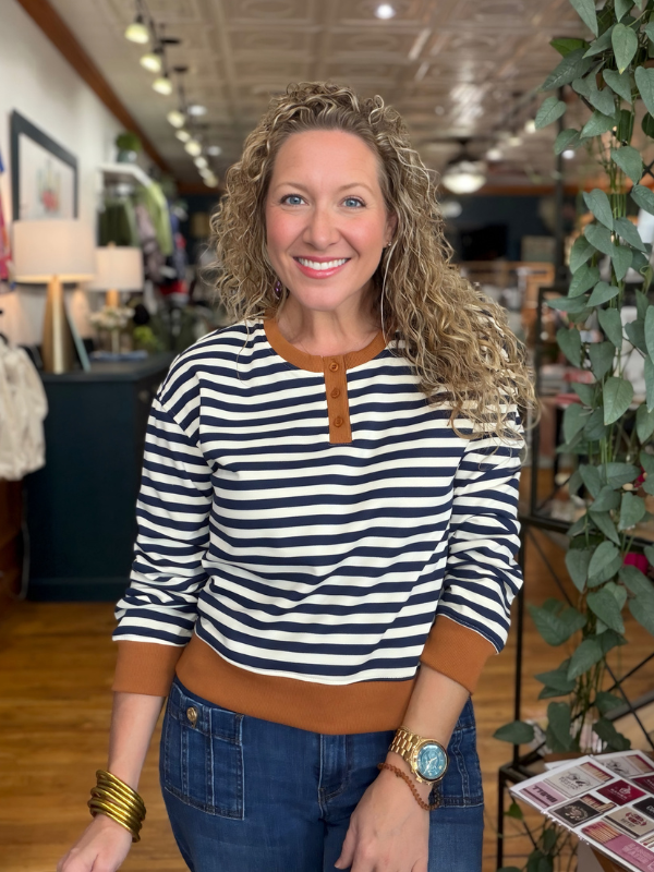 A smiling woman with curly blonde hair wearing a navy and white horizontally striped long-sleeve henley top with a brown collar and hem, paired with blue jeans, standing inside a retail store.