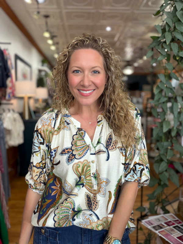 A smiling woman with curly blonde hair wearing a cream-colored short-sleeve button-up blouse with a colorful butterfly and leaf pattern, paired with blue jeans, standing inside a boutique.