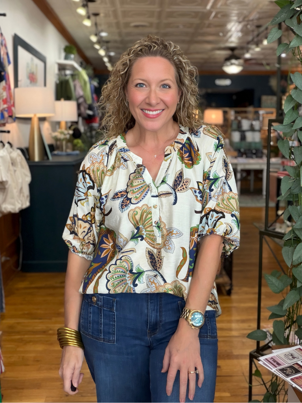 A smiling woman with curly blonde hair stands in a boutique wearing a cream-colored short-sleeve blouse with a colorful butterfly and floral print, paired with dark blue jeans.
