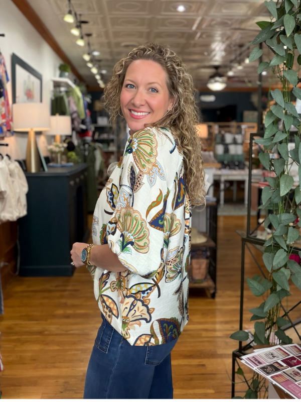 A woman with curly blonde hair stands in a boutique shop, looking back over her shoulder while wearing a cream-colored button-up blouse with a colorful tropical floral print and dark blue jeans.