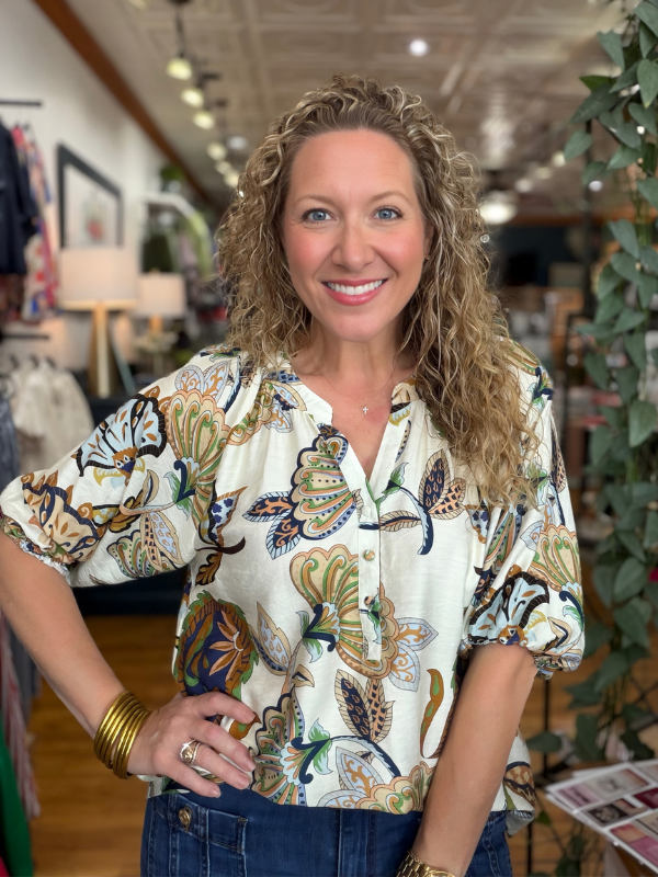 A smiling woman with curly blonde hair stands in a boutique wearing a cream-colored blouse with a colorful butterfly and floral pattern, paired with blue jeans.