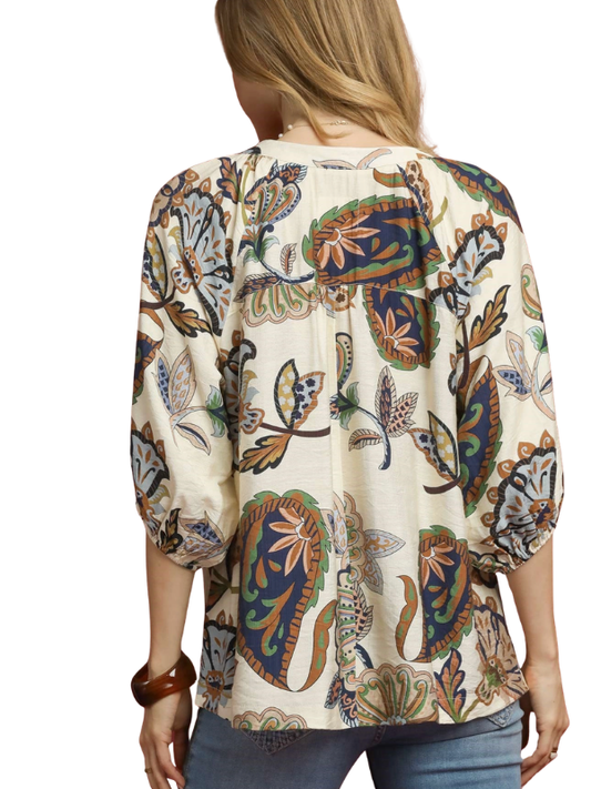 A woman viewed from the back wearing a cream-colored blouse with a detailed multicolor paisley and floral pattern, paired with blue jeans against a white background.