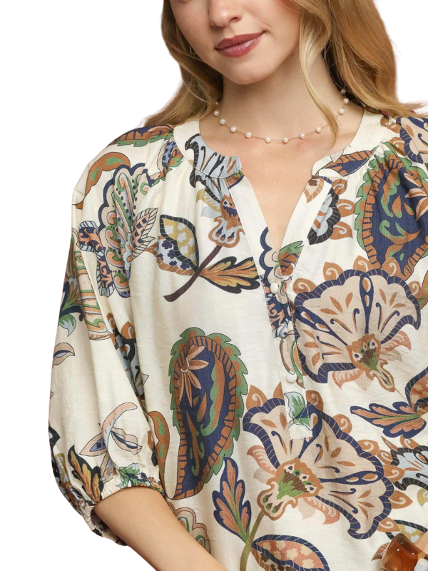 A close-up of a woman wearing a loose, short-sleeve blouse with a colorful paisley and floral print on an off-white base. The print features navy blue, brown, green, and orange motifs.
