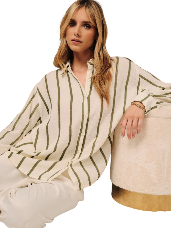 A woman wearing a cream-colored long-sleeve button-up shirt with vertical olive green stripes, paired with cream pants, sitting next to a textured cream ottoman with a gold base against a white background.